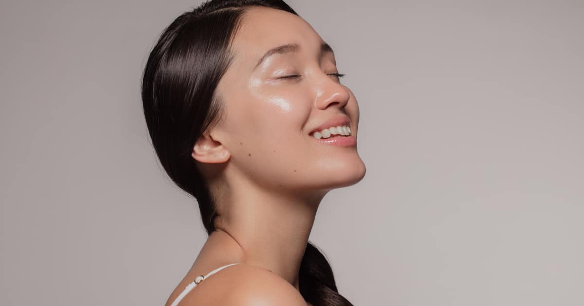 A stunning woman with smooth, radiant skin smiles while posing from a side angle.