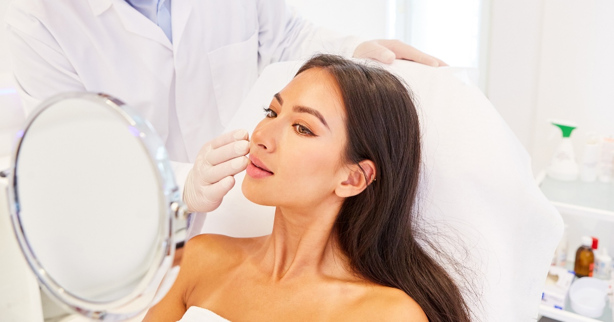 Woman seated in a cosmetic clinic chair looking into a mirror while a clinician wearing gloves gently examines or marks her cheek, with medical supplies visible in the background.